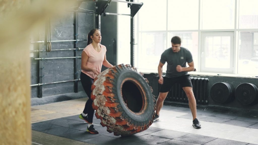 Girl in sports outfit is lifting and flipping heavy rubber tyre working out with coach in gym enjoying sports training together. People and activity concept.